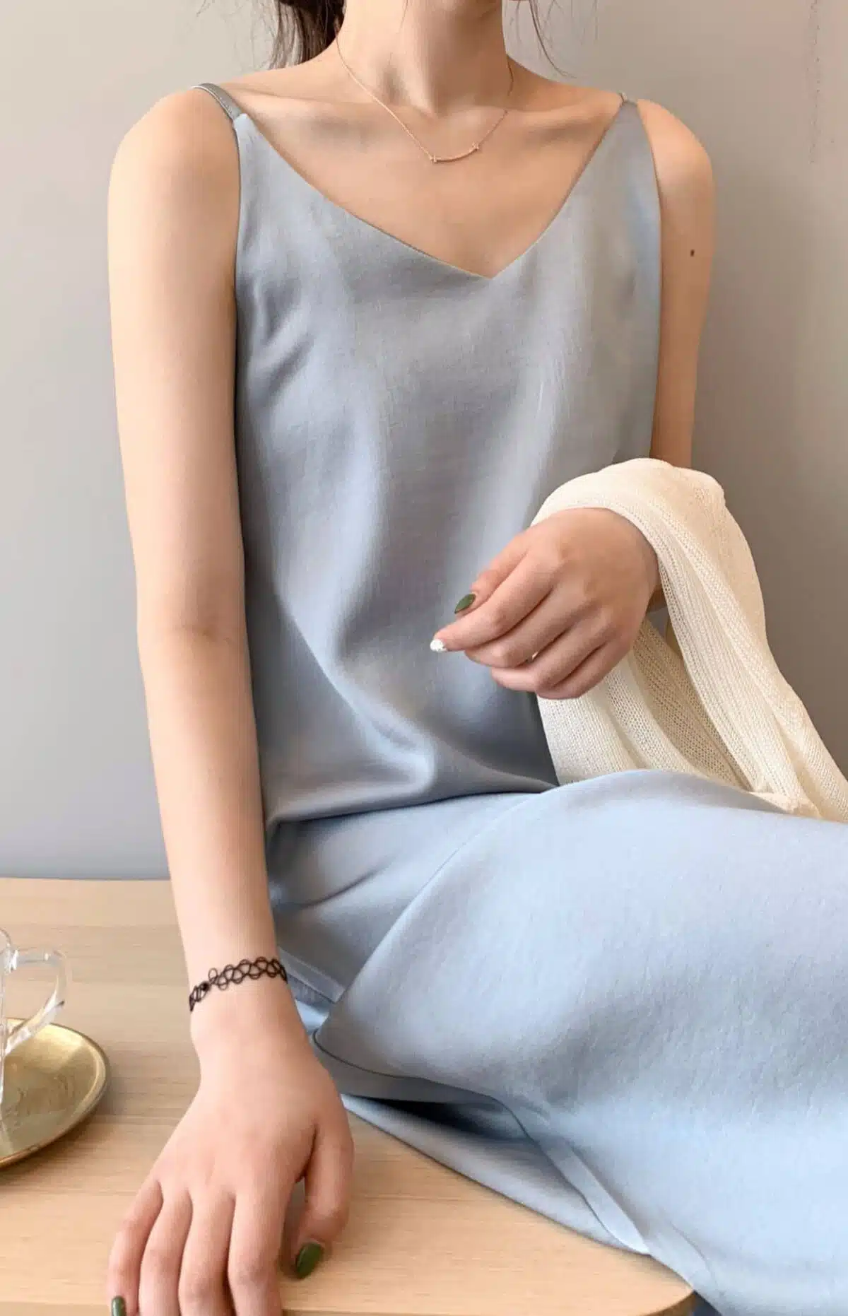 Jeune femme dont on ne voit pas la tête assise sur un banc en bois, sur lequel est posé un verre d'eau près d'elle, porte une nuissette, robe de nuit grise fluide et soyeuse à fines bretelles, et un gilet beige est posé sur son bras gauche